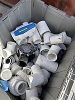 Top view of assorted white PVC pipe pieces including elbows, couplings, and valve parts inside a grey storage bin.