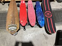Top view of 5 skateboards showing decks and logos, colors include natural wood, red, pink, blue, and black with red stripes.