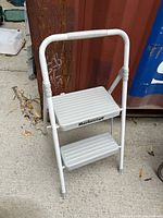 White folding 2 step metal step stool with handle positioned upright outdoors on concrete ground near a rusted metal container