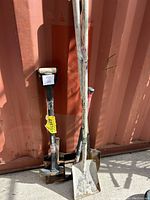 Full view of three yard tools standing upright against a container wall: pickaxe, square-edged spade shovel, digging shovel