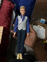 Front view of Ken doll dressed in a blue polka-dot shirt, grey vest, dark blue pants, and beige shoes held in hand against a background with boxes and furniture.