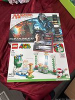 Photo of Magic The Gathering Arena of the Planeswalkers game and LEGO Super Mario Big Bill's Challenge expansion set box together on red fabric.