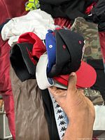 Hand holds a stack of five snapback hats showing various colors and partial embroidered logos.