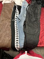 Five pairs of pants laid side by side on a bed including brown leather, black leather-style with stitching, black track pants with white logo stripe, and light blue denim jeans.