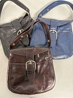 Three vintage Saks Fifth Avenue leather handbags together, showing black, blue, and dark brown colors with buckle front details and short shoulder straps with button rivets.