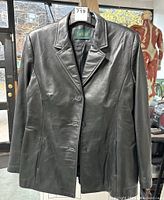 Front view of the black leather coat on hanger, showing notch lapel collar and three-button closure.