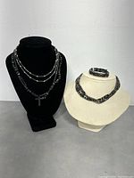 Three pieces of magnetic hematite jewelry shown on two necklace display busts: two necklaces on the black bust including one with a cross pendant, and one matching necklace and bracelet set on the cream bust.