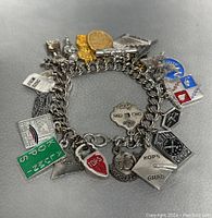 Close-up image of vintage silver-tone charm bracelet with over 20 assorted charms in varied shapes and engravings, laid flat on gray surface.