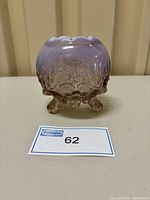 Front view of a small footed glass bowl with lavender to pink gradient and raised rose floral pattern on a plain surface.