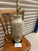 Full view of Blanc de Chine table lamp showing white ceramic carved body with floral lattice pattern, brass socket, white bulb, and black base, placed on wooden chair.