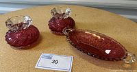 Front view of two small cranberry glass vases with clear ruffled tops and one oval cranberry glass serving dish with handles on a round table.