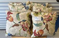 Six floral decorative pillows stacked with labels visible, displaying a colorful botanical pattern on beige fabric.