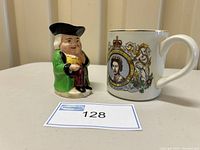Photo showing the Snuffy Toby Jug and Queen Elizabeth II mug side by side on a shelf with lot number card