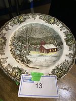 Photo of a Friendly Village dinner plate with a snowy church scene and leafy border, showing detailed design and slight wear.