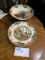 Photo of a scalloped bowl decorated with a village scene showing houses and leafless trees surrounded by green and yellow floral border pattern, set on a wooden table, along with one detailed salad plate with similar village scene and floral rim.