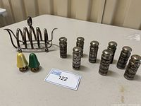 Nickel plated silver napkin holder alongside seven metal salt and pepper shakers with etched pattern and two ceramic bell-shaped shakers, displayed on a table.
