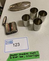 Photo of four silver-colored pewter chaudron cups, sterling silver elongated box, and silver-toned lighter on a table with labels.