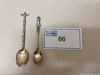 Pair of two sterling silver teaspoons with decorative handles including a crucifix and a lighthouse/tower, shown on a light surface with lot number 86 card.