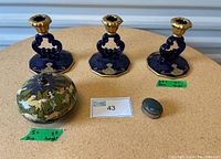 Three blue glass candle holders with gold accents, a round handcrafted trinket box with floral design, and a small covered pill box together on a round surface.