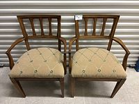 Pair of vintage wooden dining chairs with armrests and beige tufted upholstery, displayed against a white roll-up door.