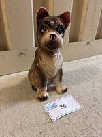 Front view of a seated ceramic dog figurine showing the facial features, ears, and front paws with label in front