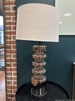 Full view of chrome mid-century modern lamp with white shade on a black round table next to brick column and dark green wall.