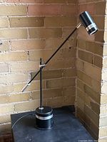 Articulated black and chrome desk lamp positioned on a black table in front of exposed brick walls.