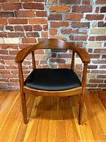 Front view of a single wood framed armchair with black vinyl seat against a brick wall
