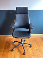 Front view of black vinyl bentwood office chair with rolling casters against a two-toned blue wall and wooden floor, showing headrest, armrests and seat upholstery.