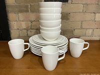 Stack of white porcelain bowls on top of plates with three coffee mugs arranged in front, displayed on wooden surface against brick wall.