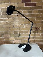 Black Workrite Ergonomics LED task light on table against brick wall, adjustable arm extended upward.