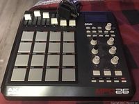 Top-down angled photo of Akai Professional MPD26 MIDI pad controller showing the layout of pads, knobs, faders, buttons, and the USB cable wrapped and placed on the device.