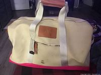 Front view of large cream and red Aria tote bag showing front zippered pocket, tan leather with embossed logo, white handles with tan leather padded grip, and tags attached. Visible dirt marks on cream fabric near bottom.