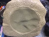 Front view of the rock-shaped Bluetooth speaker showing textured tan exterior with mesh grille in the center.