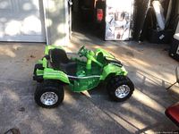 Front-side angle showing green kids buggy with large offroad tires, black seat, and steering wheel on concrete floor.