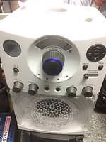 Front view of white karaoke machine showing master volume knob, control buttons, and built-in speaker grille.