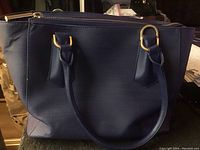 Blue purse showing front exterior with two handles attached via gold-tone rings.