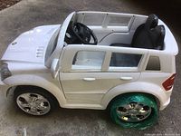 Left side view of the white Mercedes battery operated kids car showing two doors, side windows, black seats, and green plastic wrapped rear left wheel.