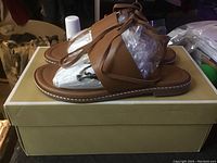 Side angle view of the brown leather Michael Kors sandals placed on a shoebox, showing ankle straps and overall shoe design.