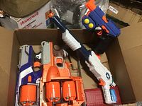 Top-down view of three Nerf blasters in cardboard box, showing different sizes and colors, including blue, orange, white, and black parts with rotating drum magazines