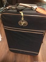 Front view of a black Pierre Cardin suitcase with brown trim, gold-tone zipper pulls, front zipped pocket, and brown handle with tags attached.