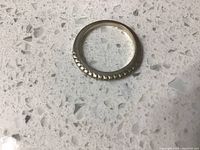 Close-up photo of silver-tone ring showing rounded band with beaded texture on surface, placed on speckled countertop.