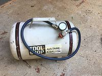 White cylindrical Tool Shop air tank with attached blue hose, pressure gauge, and valve placed on a concrete floor. Tank labeled 5 gallon capacity.