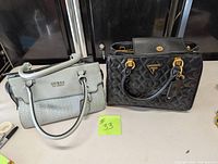 Two Guess purses side-by-side, one black quilted and one bluish grey with logo pattern