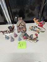 Christmas village pieces including moose house, sleighs, and trees arranged on table.