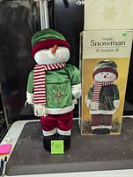 Full view of the fabric snowman greeter next to its original box showing the product branding and size.