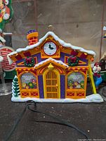 Mickey's Clock Shop figurine with snow roof, clock face, wreaths, and front door in yellow and purple colors, wired for power.