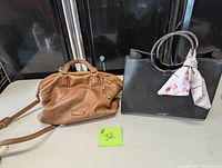 Photo showing both purses side by side, black Nine West tote on the right with floral scarf tied on handle, brown Fossil handbag on the left with key charm and long shoulder strap