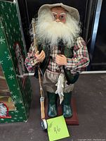 Front view of the fly fishing Santa figurine holding a fishing rod and fish with the green box behind it.