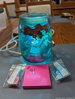 Photo of blue glass Scensty Little Mermaid electric wax warmer burner with red-haired mermaid illustration, placed on a wooden table with two small sealed packets of scented wax melts in front labeled with a pink sticky note '#91'.
